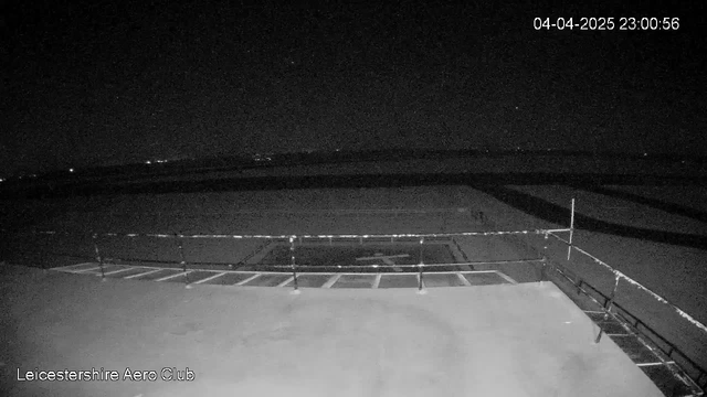 A dark, nighttime scene captured by a webcam at the Leicestershire Aero Club. In the foreground, there is a flat rooftop with a railing. Beyond the rooftop, an expansive area appears mostly empty, with a few distant lights visible in the dark, suggesting a landscape that may include hills or structures. The image reflects a clear sky with some stars. The date and time are displayed in the top right corner, indicating April 4, 2025, at 11:00 PM.