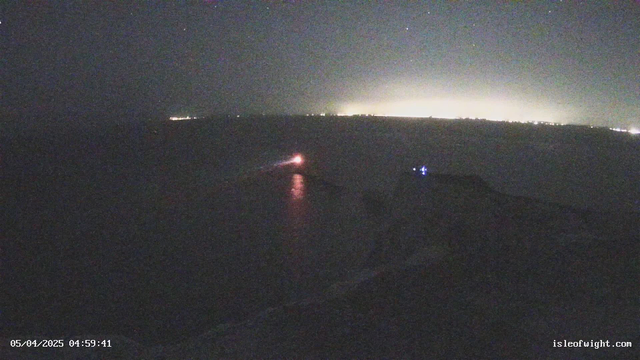 The image shows a nighttime coastal scene. In the foreground, there is a dark shoreline with the water reflecting some light. A small boat with a visible light is navigating near the shoreline. In the background, faint lights from distant buildings or a city create a glowing horizon across the water. The scene is mostly dark, but the lights provide some contrast against the night sky.
