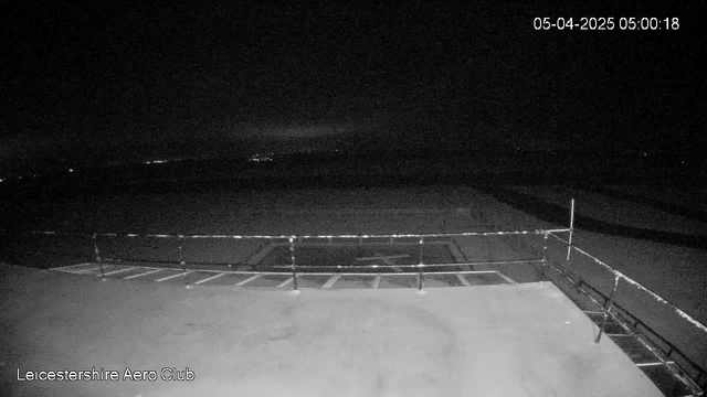 A dimly lit scene shows a flat rooftop with a railing on the left side. In the distance, faint lights from buildings or vehicles are visible against a dark sky. The image appears to be taken at dawn or in the early morning hours, with minimal visibility. The date and time are displayed in the upper right corner.
