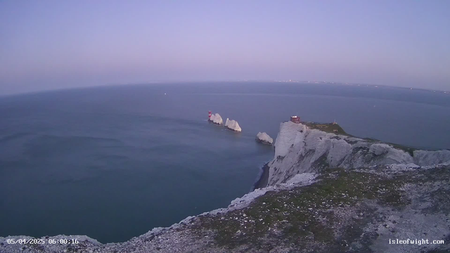 A coastal view featuring white chalk cliffs extending into the ocean, with a series of rocky formations in the water. Two lighthouses, one red and one at the base of the cliffs, are visible among the rocks. The sky is a gradient of soft colors, transitioning from light blue at the horizon to pale lavender overhead. The ocean appears calm, reflecting the colors of the sky. There are a few distant ships and faint lights visible in the background.