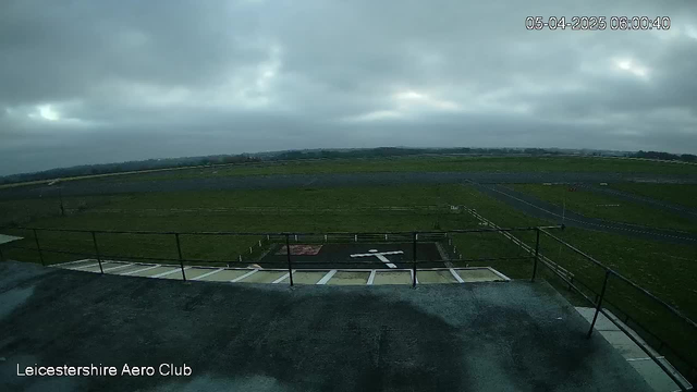 A view from a webcam positioned on a building at the Leicestershire Aero Club, showing an expansive grass airfield under overcast skies. The foreground includes a flat rooftop and a railing, along with a helipad marked with a large white cross and a red square. In the background, the airfield extends towards a distant tree line, with multiple taxiways visible across the landscape. The image suggests early morning light with a cool, muted atmosphere.