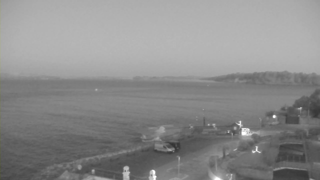 A coastal scene at dusk, presented in black and white. The foreground features a parking area with a vehicle parked near the edge of a stone wall. In the background, the water appears calm with gentle waves and hints of light reflecting off its surface. A distant shoreline with trees and hills fades into the foggy atmosphere. The sky above transitions to a lighter shade, suggesting twilight.