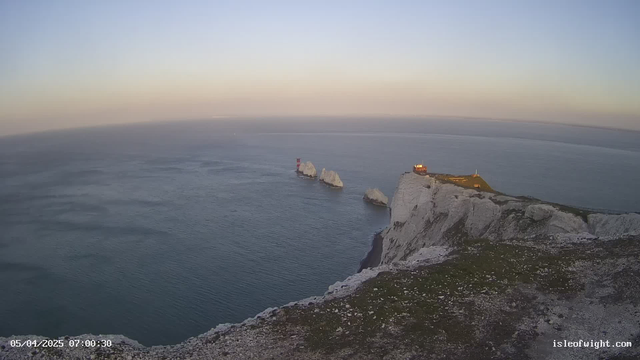 A panoramic view of the sea at dawn, with gentle waves reflecting soft sunlight. In the foreground, a rocky cliff with green grass leads to steep white chalk cliffs. Offshore, several rocky formations emerge from the water, with one featuring a red and white striped lighthouse. A small building sits on top of the cliffs, illuminated in warm light, while the sky transitions from blue to pastel hues near the horizon.