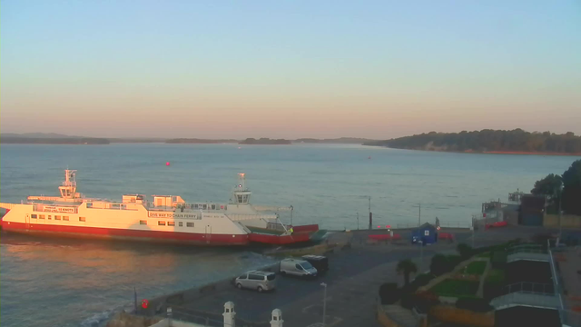 A ferry is docked at a pier on a calm body of water during sunset. The ferry is mostly white with a red lower hull and has text reading "GIVE WAY TO CHAIN FERRY." In the foreground, there are parked vehicles. The background features a gentle gradient of orange and blue in the sky, with silhouettes of hills and trees on the horizon. The water is mostly still, reflecting the colors of the sky.