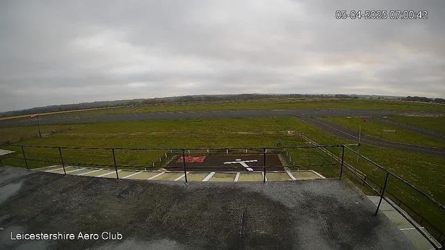 A view from a webcam overlooking a grassy airfield. The sky is overcast with gray clouds. In the foreground, there is a flat rooftop area, followed by a railing. The airfield features a large, painted white "X" on the ground, indicating a landing area, and there are scattered sections of neatly maintained grass. A road runs alongside the airfield, with a few wooden fences visible in the distance. The image timestamp indicates it was taken early in the morning.