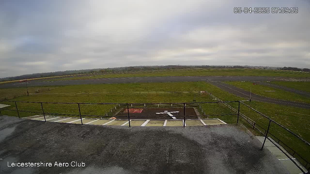 A wide view of a grassy airfield under a cloudy sky. In the foreground, there is a wooden railing with a ramp leading down. The airfield is empty, with a runway extending into the distance. A white cross is painted on the ground, next to a red-painted area. The date and time, April 5, 2025 at 07:59:53, are displayed in the top right corner.