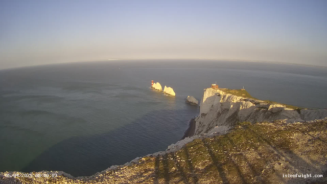 A panoramic view of a coastal scene featuring white chalk cliffs and the ocean. In the foreground, the cliff edge is visible with scattered rocks and grass. Out in the water, there are two white rock formations with a red-and-white striped lighthouse on one of them. The sky is clear with a light blue hue, and the ocean appears calm, reflecting the sunlight.