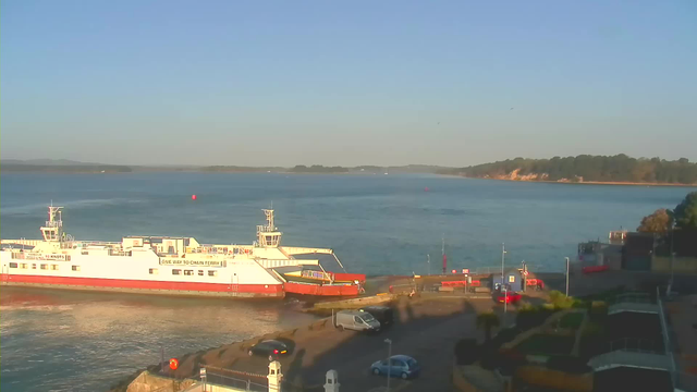A ferry is docked at a harbor near the water, with a clear blue sky overhead. The ferry is mostly white with some red accents and a sign indicating "Give way to chain ferry." In the foreground, there are parked cars and a paved area, while a shoreline with trees and cliffs is visible in the background. The water appears calm, reflecting the sky's colors.