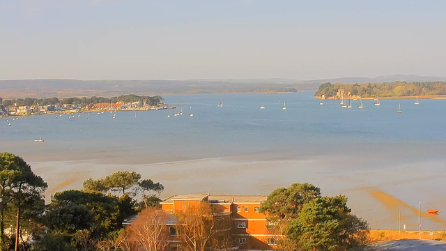 A serene view of a blue body of water, with several small boats scattered across it. In the background, a calm shoreline dotted with trees and buildings can be seen. On the far side of the water, a gentle rise of land features a small castle or historical structure. The sky is clear, with soft sunlight reflecting on the water's surface.