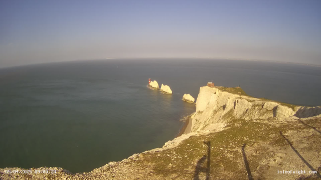 A coastal view featuring rugged white chalk cliffs that drop into a calm blue sea. In the distance, there are two prominent white rock formations with a red lighthouse on one of them. The horizon is mostly clear under a blue sky, with a few wispy clouds. The foreground shows the edge of the cliff with patches of green grass and some rocky terrain. The image is taken during daylight, highlighting the natural landscape.