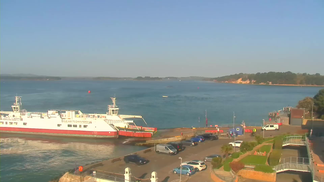 A calm body of water reflects the clear blue sky, with a white ferry docked at a pier on the left side of the image. The ferry has red accents and is situated next to a parking area filled with various cars. In the background, there are green trees and a sandy shoreline. Small boats can be seen on the water, and there are several buoys marking the channel. The scene is tranquil, indicating a sunny day.