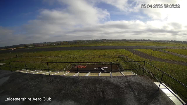 A panoramic view of a grassy airfield under a partly cloudy sky. The foreground features a flat rooftop with a railing. In the middle ground, an airstrip runs horizontally, bordered by a green field with a white cross marking a landing area. There is a wooden fence surrounding the grassy field, and the horizon is lined with a low, distant landscape. The timestamp on the image shows it was taken on April 5, 2025, at 9:00 AM.