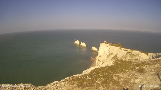 A scenic coastal view featuring white chalk cliffs meeting the calm blue sea. In the foreground, the cliff edge is rocky and grassy, while further out, two distinct rock formations rise from the water, each topped with a small red and white lighthouse. The sky is mostly clear with a slight gradient of blue, indicating a sunny day.