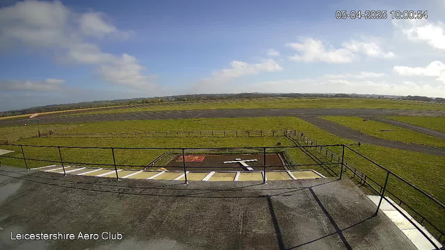 A wide view of a grassy airfield under a clear blue sky with scattered clouds. In the foreground, there is a low railing and a flat surface. A white cross is visible on a brown patch of ground, suggesting a marked area. The airfield features a runway, bordered by a wooden fence, with several grassy areas around it. A windsock is visible, indicating wind direction, and a tarmac road can be seen in the distance leading away from the airfield. The date and time are displayed in the top right corner.