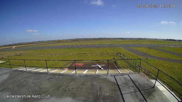 A clear blue sky with a few scattered clouds is visible overhead. Below, a grassy area with a fenced boundary stretches out, leading to an airstrip. The airstrip is paved and extends into the distance, with a ramp-like path. In the foreground, there is a helipad marked with a white cross on a dark surface, accompanied by a red square nearby. The overall scene conveys a calm, sunny day at an aerodrome.