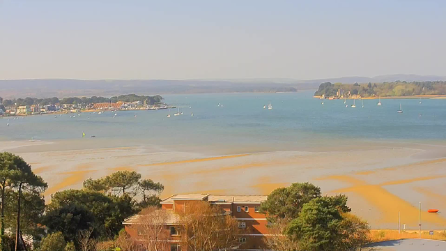A panoramic view of a serene coastal scene. In the foreground, there are patches of sandy shore with some greenery and trees. The water stretches out towards the horizon, where several small boats can be seen sailing. To the left, a picturesque seaside town with colorful buildings lines the shore. In the distance, gentle hills create a backdrop against the clear blue sky. The sunlight casts a warm glow over the entire scene.