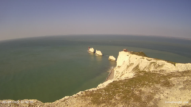 A panoramic view of a coastline featuring steep, white chalk cliffs rising above a calm sea. In the distance, three sea stacks are visible, with a red lighthouse on the first stack. The sky is clear with a slight gradient of blue. The foreground includes grassy terrain and a sandy beach along the water’s edge. The scene captures a tranquil, sunny day at the seaside.