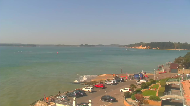 A sunny coastal scene featuring a calm sea with shades of blue and green. In the foreground, there is a parking area with several parked cars, including white, red, and gray vehicles. People are walking near the water's edge, and a few small boats can be seen in the distance. The shoreline is lined with trees and a sandy beach area, while a concrete pier extends into the water. The sky is clear with minimal clouds.