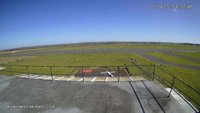 A clear blue sky is visible above a large green field. In the foreground, there is a flat rooftop with a railing. Below the rooftop, a marked area shows a white shape resembling a small airplane, set against a dark surface. Several taxiways and a runway can be seen extending across the grassy area. The overall scene depicts an aerial view of an airfield, with an empty landscape suggesting a calm day. The current date and time are displayed on the top right corner.