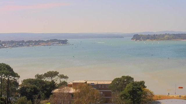 A serene view of a bay with calm blue waters stretching into the distance. Small boats and sailboats are scattered across the water, while a few kite surfers can be seen in the foreground. Lush trees and shrubs frame the lower part of the scene, with a building visible in the foreground. In the background, gentle hills rise against a clear sky.