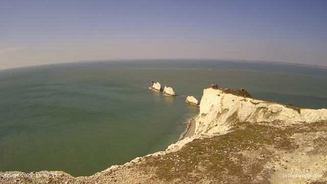 A scenic view of a coastal area featuring white cliffs and a turquoise sea. In the foreground, there is a rocky shoreline with greenery. In the background, several white rock formations rise from the water, with one small red structure visible on one of the rocks. The sky is clear and blue, creating a tranquil atmosphere.