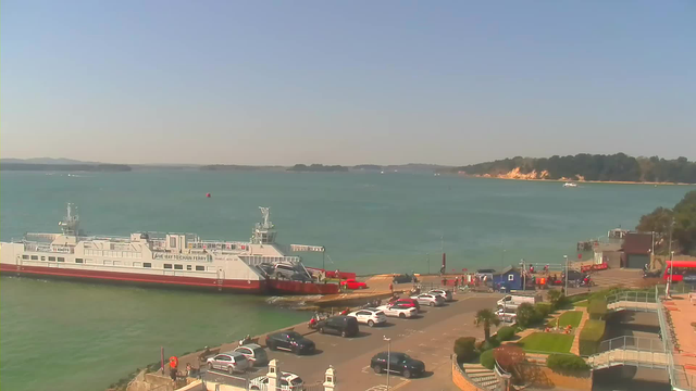 A ferry is docked at a pier with a sandy beach and cliffs in the background. The water is turquoise blue, and there are several boats in the distance. The foreground features a parking area with various cars and people walking. Lush green plants are visible along the walkway, and a small building is nearby. The sky is clear with few clouds.