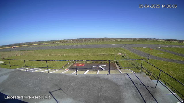 A clear sky with no clouds is visible above a grassy landscape. In the foreground, there is a flat rooftop with a low railing. Below, a runway stretches across the landscape, leading to a grass field bordered by a white fence. In the bottom left corner, there is a helicopter landing pad marked with a white figure. The scene is well-lit, suggesting it is daytime.