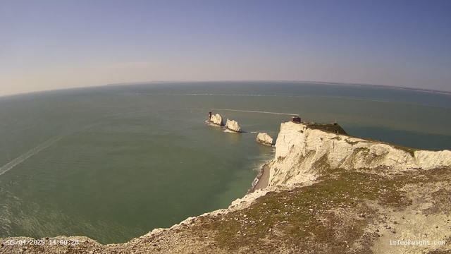 A panoramic view of a coastal landscape featuring bright blue skies and calm, greenish-blue waters. In the foreground, there are white chalk cliffs with some vegetation. Several rocky outcrops emerge from the water, one of which has a small structure on top. The horizon is distant, with a few small boats visible leaving trails in the water. The overall scene is serene and scenic, showcasing nature's beauty.