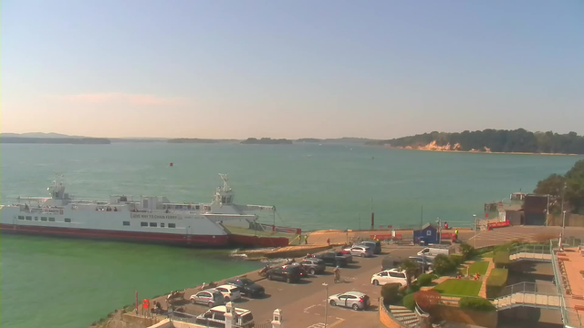 A seaside view featuring a large white ferry docked at a terminal, surrounded by calm green water. In the foreground, there is a parking area with several parked cars and some people walking. The background shows a coastline with greenery and a sandy area, under a clear blue sky.