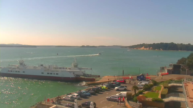 A clear, sunny view of a marina featuring a large ferry docked at the side. The ferry is white with red accents. In the foreground, there is a parking lot filled with various cars, predominantly white, red, and yellow. People can be seen walking around the area. The water is calm with a turquoise hue, and there are several small boats on the water. In the background, there are rolling hills and a sandy beach. The sky is bright blue with no clouds.
