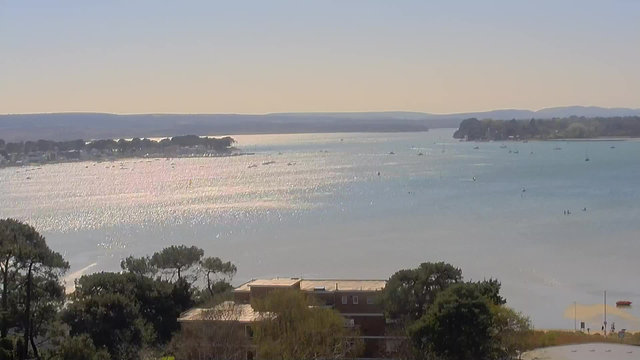 A serene waterfront scene features shimmering water reflecting sunlight, with multiple small boats scattered across the bay. In the background, a gentle shoreline lined with trees and buildings meets the water, while rolling hills are visible in the distance under a clear blue sky. Near the foreground, a mixture of greenery and structures adds depth to the landscape.