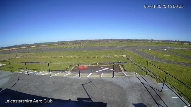 A clear blue sky stretches overhead, with no clouds visible. In the foreground, there is a flat rooftop railing with a shadow extending across the surface. Below, an open field is visible, bordered by a wooden fence. The grass is green and well-maintained, while a large, empty runway runs horizontally across the midsection of the image. A section of the surface has a red marking and a white 'X' on it, indicating a helipad or similar landing area, while several signs and markings can be seen along the sides of the runway. The overall scene conveys a peaceful, sunny day at an airfield.