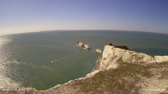A panoramic view of a coastal landscape featuring white chalk cliffs meeting the sea. In the foreground, the cliffs are rugged with patches of green vegetation. The ocean is visible in shades of blue and green, reflecting sunlight, with a small boat leaving a wake as it moves through the water. The sky is clear and bright blue, and a distant landmass can be seen on the horizon.