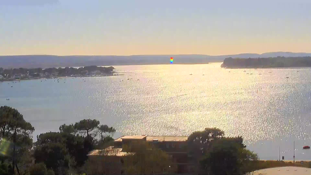 A serene waterfront scene showing a wide body of shimmering water reflecting sunlight. In the background, soft hills rise against a clear blue sky, with a few small boats scattered across the water. On the shore, there are trees and buildings partially obscured by foliage. The overall atmosphere conveys a peaceful and tranquil environment.