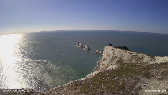 A panoramic view of a coastline under a clear blue sky. The scene features steep white cliffs alongside a calm sea, which reflects sunlight, creating a shimmering effect. In the water, distinctive rock formations can be seen jutting out, with one rock having a small structure on top. The shoreline is composed of a mix of sand and gravel, framed by grass-covered cliff tops.