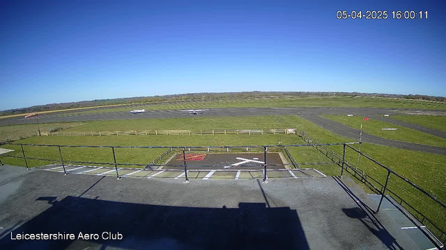 A clear blue sky is visible above the Leicestershire Aero Club, with bright sunlight illuminating the scene. In the foreground, there is a flat rooftop structure with a railing. At the center of the image, a helipad marked with a red square and white cross is seen. Below the helipad, a grassy area leads to a runway, where a small airplane is preparing for takeoff. A wind sock is positioned to the left, indicating the direction of the wind. The far background features a neatly maintained grassy field with distant hills beyond the runway.