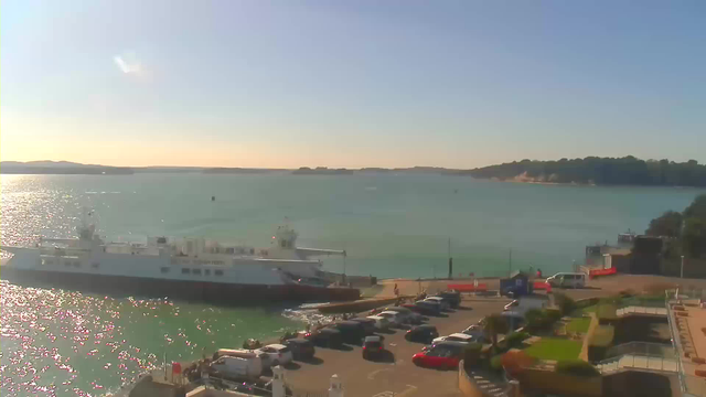 A bright landscape featuring a calm body of water reflecting sunlight. In the foreground, a large white ferry is docked at a port, with several parked cars lining the shore. The area includes a small grassy patch and some low shrubs. In the background, there are distant hills and trees under a clear blue sky.