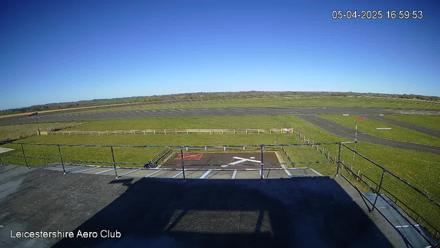 A wide view from a webcam at Leicestershire Aero Club shows a clear blue sky above a grassy airfield. In the foreground, there is a flat rooftop with a shadow, and a white cross marking on the ground. In the distance, the airfield is visible with several runway paths intersecting, bordered by a wooden fence. A wind sock stands on the left side of the image, indicating wind direction. The date and time are displayed in the upper right corner.