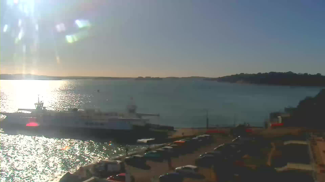The image shows a bright sunny day at a waterfront location. In the foreground, there is a ferry docked at a terminal, and the water is sparkling in the sunlight. Several cars are parked along a road near the waterfront, and the horizon features a distant view of a tree-covered landmass. The sky is clear with a gradient of light blue, indicating a calm and pleasant atmosphere.