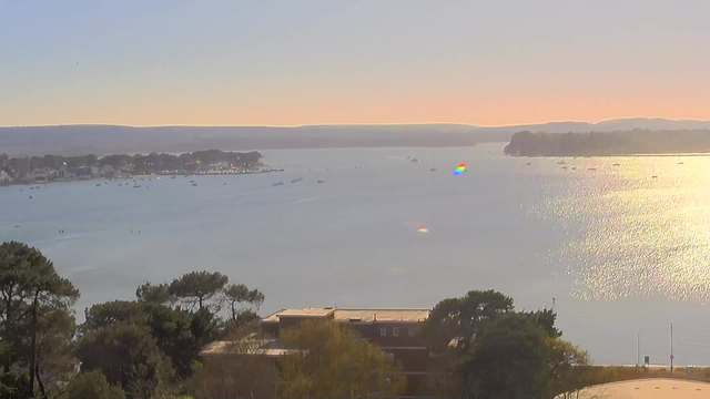 A serene water view captures a bay with calm blue waters reflecting sunlight. Numerous small boats are scattered across the water, and a faint rainbow shimmer is visible on the surface. In the background, hills rise under a clear sky, with a slight gradient from blue to soft orange near the horizon. Trees and buildings frame the foreground, adding depth to the landscape.