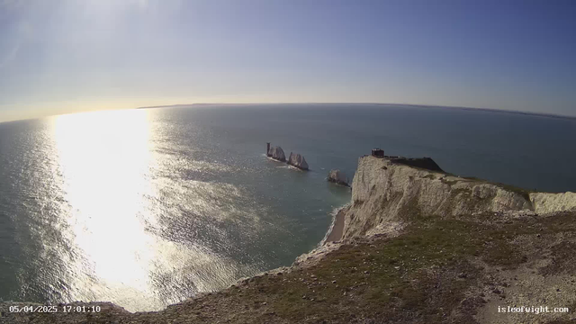 A view from a high cliff overlooking the ocean, reflecting bright sunlight. In the distance, several rock formations rise from the water. A small building is visible at the edge of the cliff to the right. The sky is clear with a few wispy clouds, blending into the blue horizon. The foreground features grassy terrain with scattered stones.