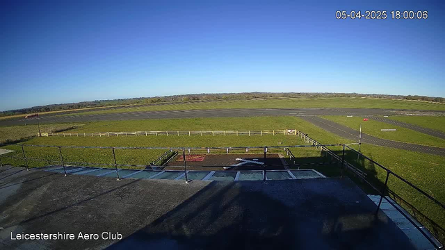 A clear blue sky is visible above a grassy airfield with several runways. In the foreground, there is a fenced area and a building rooftop with a railing. The scene is well-lit, and the horizon shows distant trees. The date and time display indicates it is April 5, 2025, at 6:00 PM.