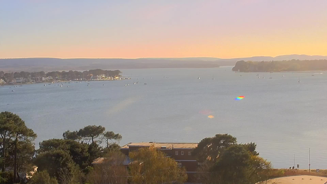 A serene view of a body of water under a soft pastel sky, transitioning from light blue to shades of orange and yellow at the horizon. The calm water reflects the colors of the sky. In the distance, small boats are scattered, with a few on the left side near a shoreline dotted with buildings and trees. The foreground features lush green trees, adding depth to the scene. Overall, the image conveys a peaceful, tranquil landscape.