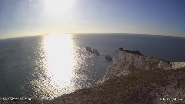 A panoramic view of a coastline featuring steep white cliffs on the right, with bright sunlight reflecting off the calm ocean waters. In the distance, several jagged rock formations are partially submerged in the sea. The sky is clear and blue, with a few wispy clouds visible. The scene conveys a tranquil and scenic environment.