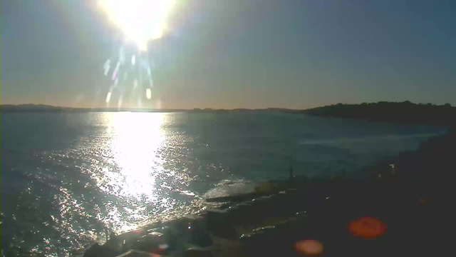 The image captures a scenic view of a body of water reflecting bright sunlight. The sun is positioned in the upper left corner, casting a glare and illuminating the surface of the water, creating sparkling highlights. In the background, there are distant hills or landforms silhouetted against the horizon. The foreground shows the water gently lapping at a rocky shoreline, with some visible structures and vehicles partially illuminated. The overall atmosphere is calm and tranquil, typical of a clear day outdoors.