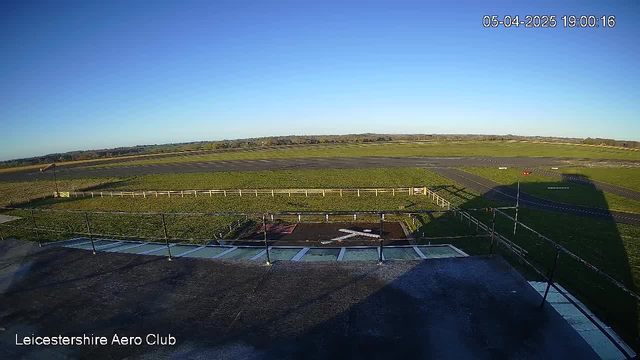 A wide-angle view from a webcam at the Leicestershire Aero Club shows an open grassy field and a runway. The sky is clear and blue, with no clouds. In the foreground, there is a fenced area that contains a marked spot on the ground resembling an "X." The edge of a roof is visible at the bottom, with several panels and a railing. In the distance, the horizon consists of trees and a sprawling landscape. The image indicates the time as 19:00:16 on April 5, 2025.