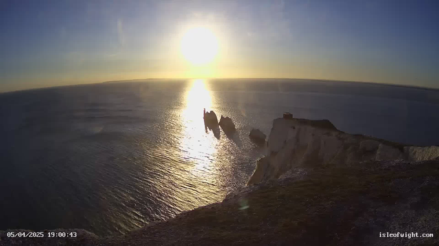 A scenic view of the sea during sunset, with the sun low on the horizon illuminating the water. The surface of the ocean reflects the golden light, creating a shimmering effect. In the foreground, there are rugged cliffs, with white rock formations jutting into the water. Several large rocks stand in the ocean, creating an interesting silhouette against the sunset. The sky transitions from blue to warm hues of orange and yellow as the day ends. In the distance, a faint outline of land can be seen on the horizon.