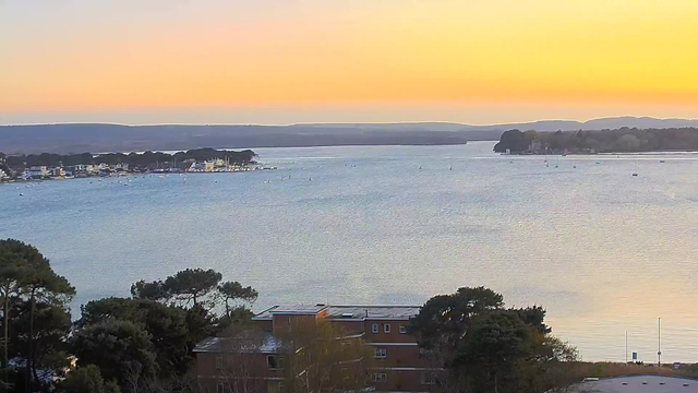 A serene view of a body of water during sunset, with soft orange and blue hues in the sky. The water reflects the colors of the sunset, creating a calm atmosphere. In the foreground, there are trees, and further back, buildings line the waterfront. Several small boats are visible on the water, and hills can be seen in the distance. The overall scene conveys tranquility and natural beauty.