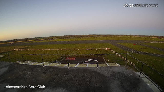 A wide view of an outdoor scene captured by a webcam, featuring a grassy airfield under a clear sky at dusk. In the foreground, there is a metal railing indicating a balcony area, along with a paved landing strip marked with a white "X" symbol on black asphalt. Beyond the landing area, an expansive green grass field extends towards a distant horizon, with a few scattered trees and a light breeze indicated by a windsock. The scene is tranquil and devoid of aircraft, suggesting a quiet time at the airfield.