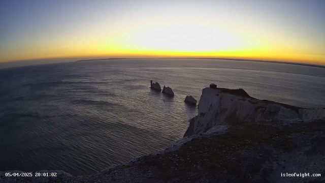 A coastal scene at sunset, featuring a view of the ocean and a series of prominent white chalk cliffs on the right side. In the foreground, the cliffs slope down towards the water. Three distinct rock formations jut out from the sea, with the furthest rock displaying a small tower or structure on it. The sky transitions from orange near the horizon to a lighter blue above, indicating the fading light of dusk. The water appears calm with gentle ripples reflecting the colors of the sky.